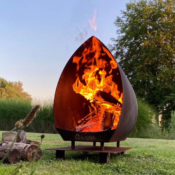 Coretnstaal tuinhaard met vlammen, hout en afgebeeld in het gras met een boom op de achtergrond